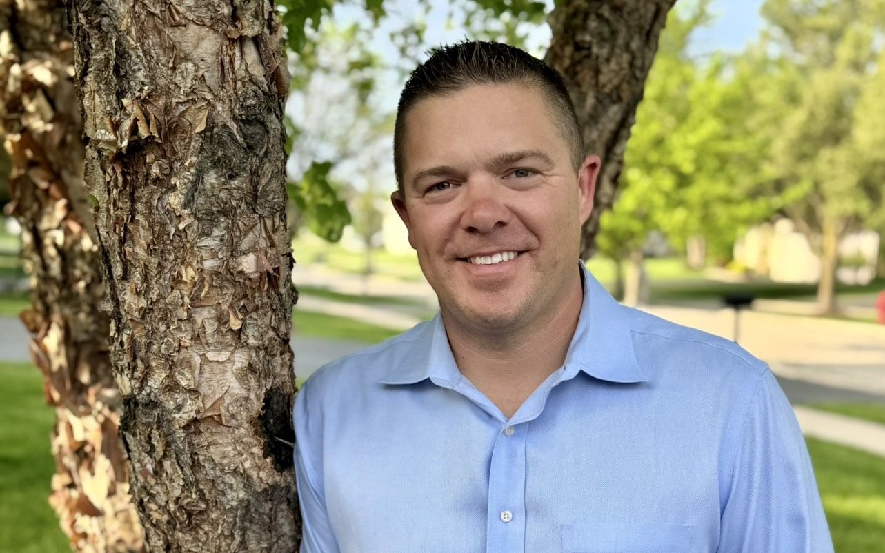 A man in a light blue dress shirt smiles while standing next to a tree. The background features a suburban street, green grass, and leafy trees on a sunny day.