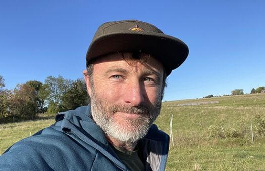 Joe, outdoors on a sunny day, standing in a field with trees in the background. Wearing a blue jacket and are looking slightly off camera with a neutral expression.