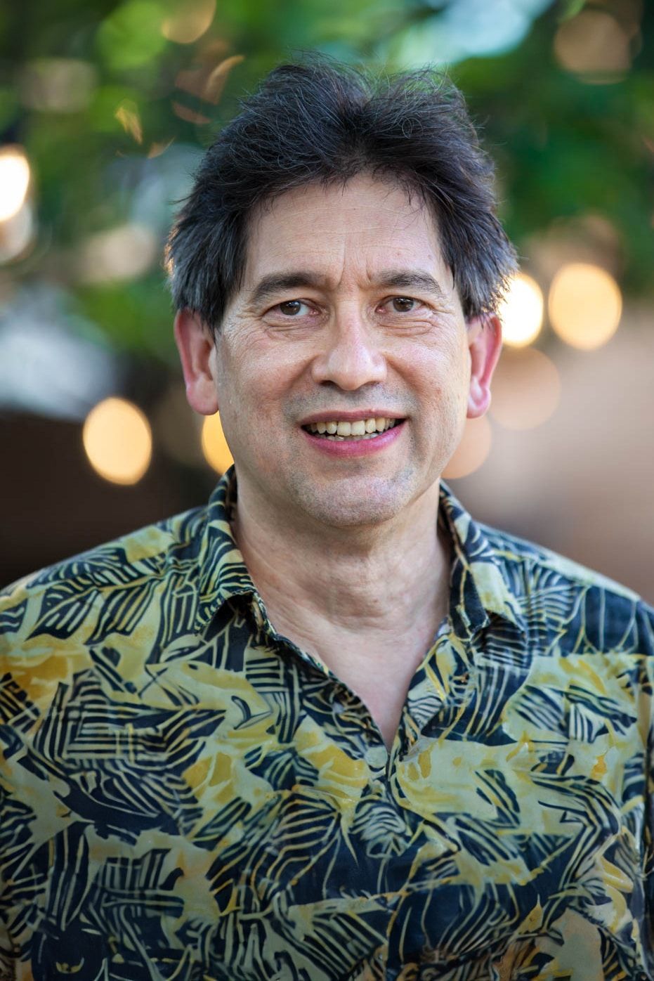 A smiling man with dark hair stands outdoors, wearing a patterned shirt with leaves. The background is blurred with bokeh from sunlight filtering through green foliage.