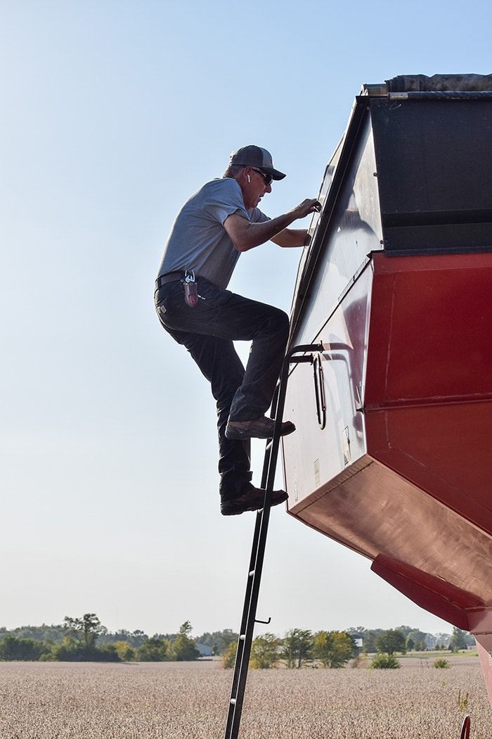 A man wearing a cap and sunglasses climbs a ladder attached to a large red agricultural machine in a field under a clear sky.