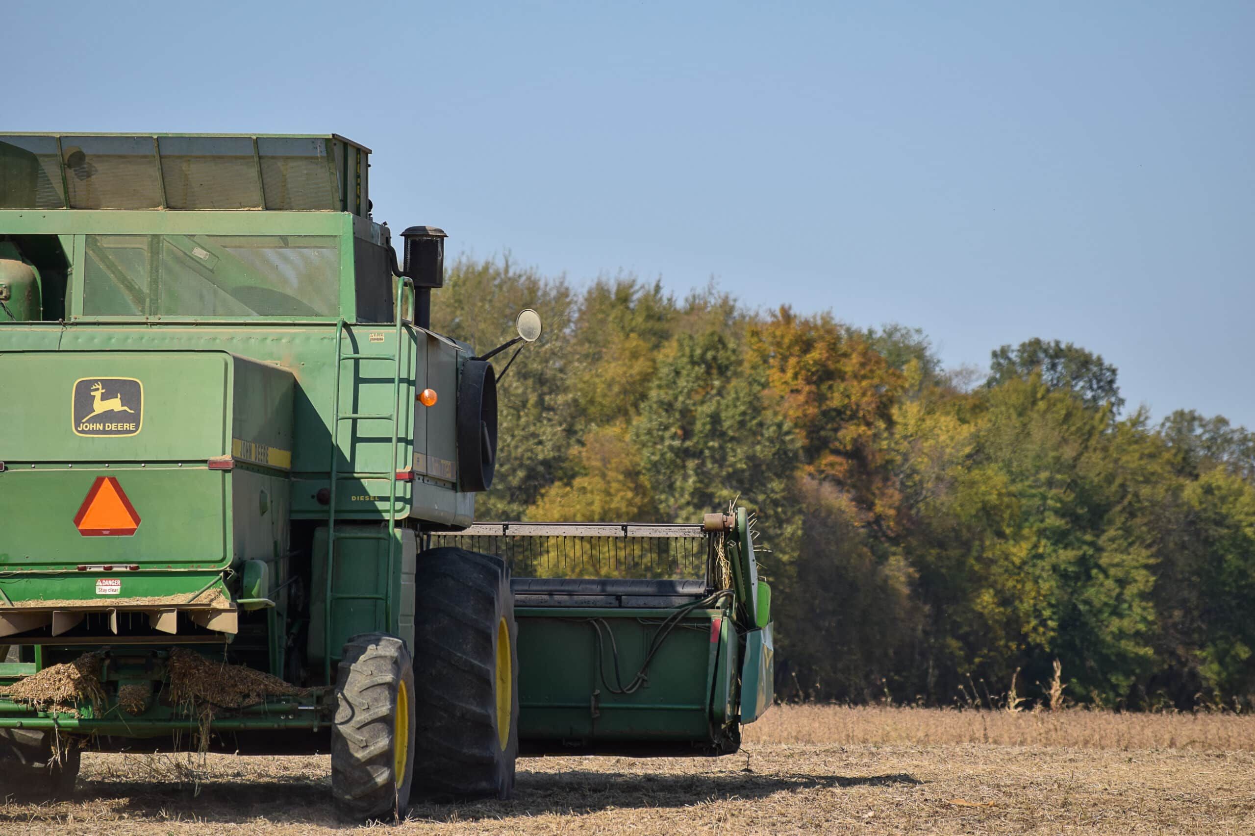 A green John Deere combine harvester sits stationary in a harvested field, with trees showing early autumn colors in the background under a clear blue sky.