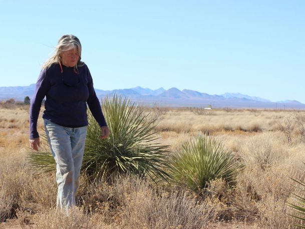 A woman walks through a dry, grassy desert landscape with distant mountains under a clear blue sky. She wears a dark long-sleeve shirt and jeans, with sunglasses hanging from her neckline. Surrounding vegetation includes sparse shrubs and yucca plants.
