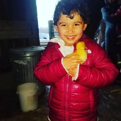 A young child wearing a bright pink jacket smiles while holding a small yellow duckling indoors, with a trash can and other people visible in the background.