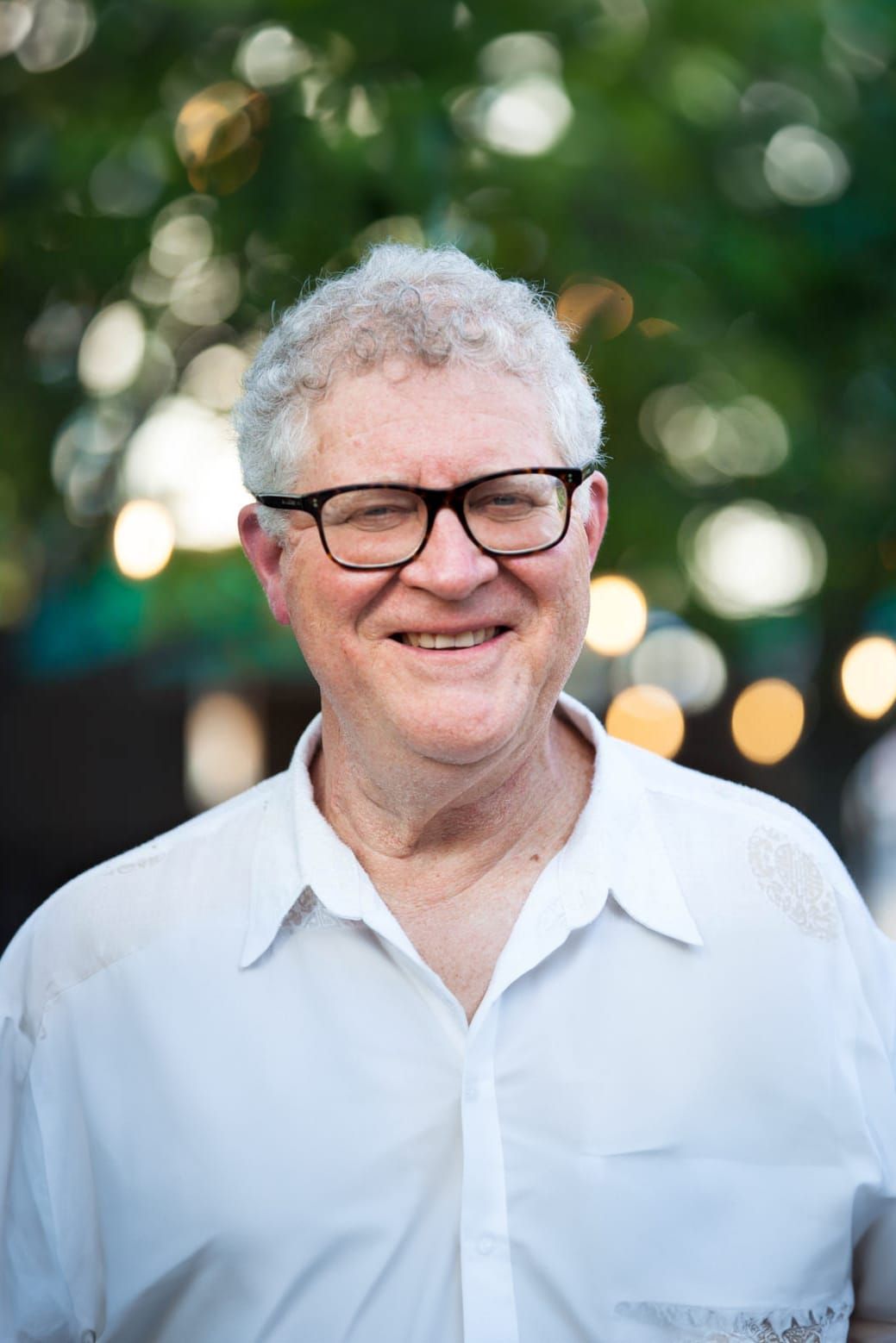 Smiling older man with glasses and curly white hair stands outdoors wearing a white shirt. The background is blurred with green foliage and circular light bokeh.