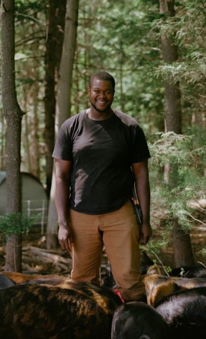 A veteran farmer in a black shirt and tan pants stands smiling in a wooded area with several pigs at his feet. Trees and a metal shelter are visible in the background as sunlight filters through the foliage.