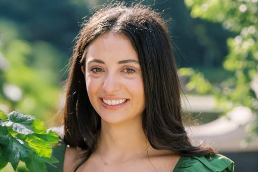 A young woman with long dark hair smiles outdoors, surrounded by green leaves and sunlight. She wears a green top and a delicate gold necklace. The background is blurred with natural greenery.