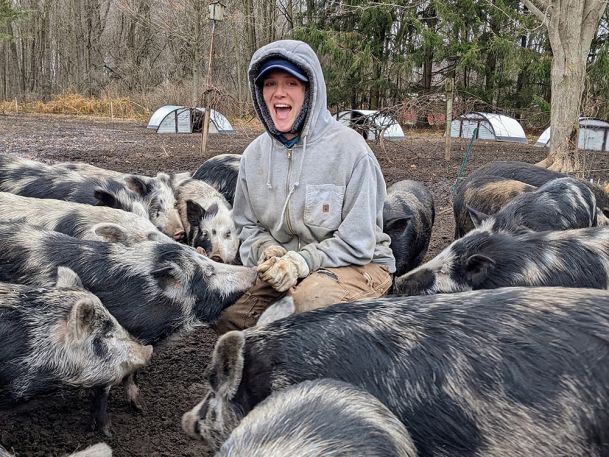 A person in a gray hoodie and gloves kneels among a group of black and white pigs in an outdoor, muddy area. They are smiling and surrounded by trees and small structures in the background.