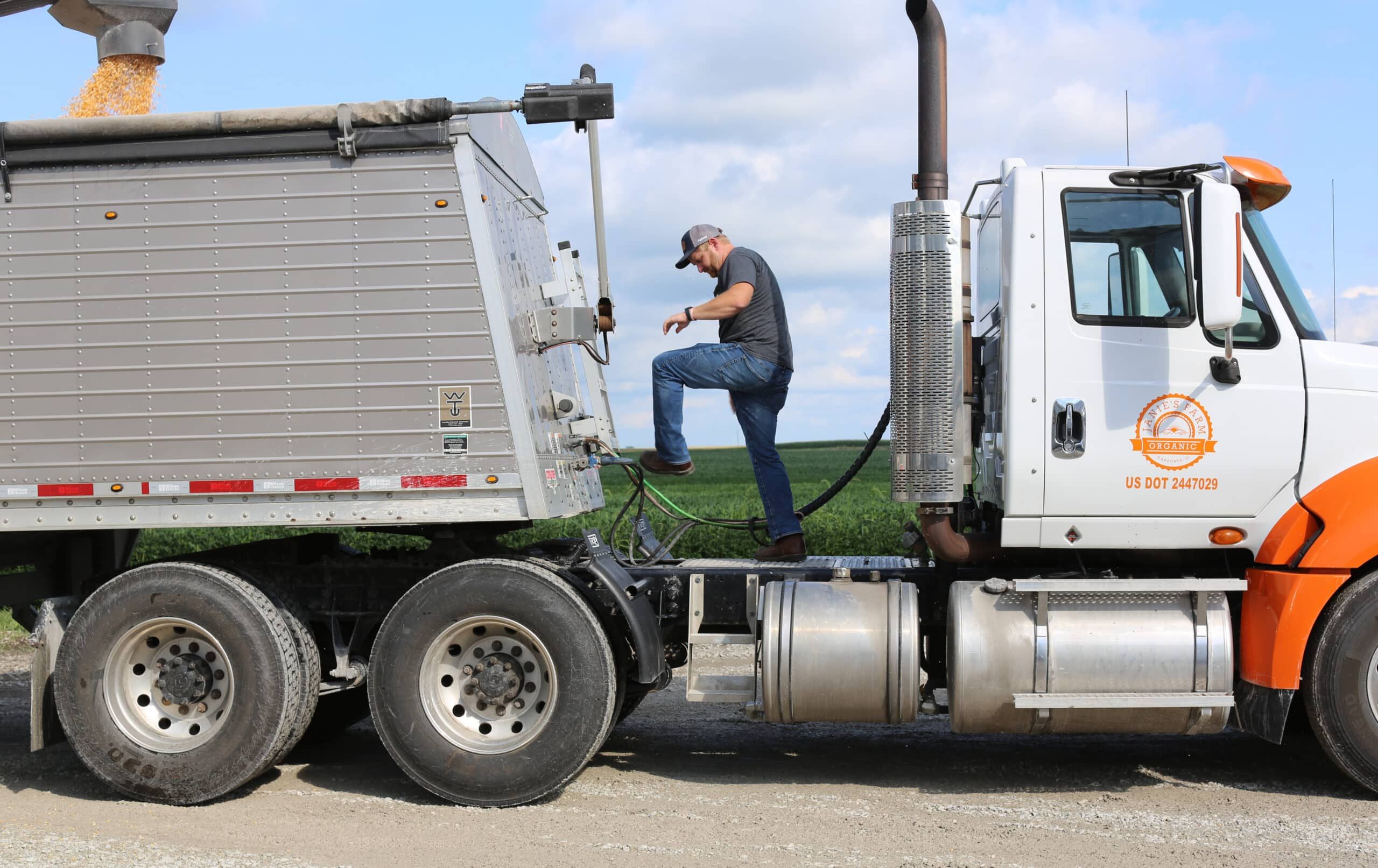 A man wearing a cap and jeans steps onto a platform between a white and orange semi-truck and a silver grain trailer, with corn pouring into the trailer. Green fields and a partly cloudy sky are in the background.