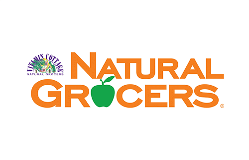 Logo for Vitamin Cottage Natural Grocers: the text "Natural Grocers" in orange, with a green apple shape as the "O." A smaller sign reads "Vitamin Cottage Natural Grocers" with a purple and green design.