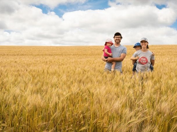 A family of four stands in a golden wheat field under a blue sky with fluffy clouds. The man holds a young child in pink, while the woman holds an older child wearing a cap. They all wear casual clothes and smile at the camera.