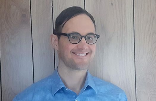 Matt, smiling while wearing a blue checkered shirt. Standing in front of a wooden panel wall.