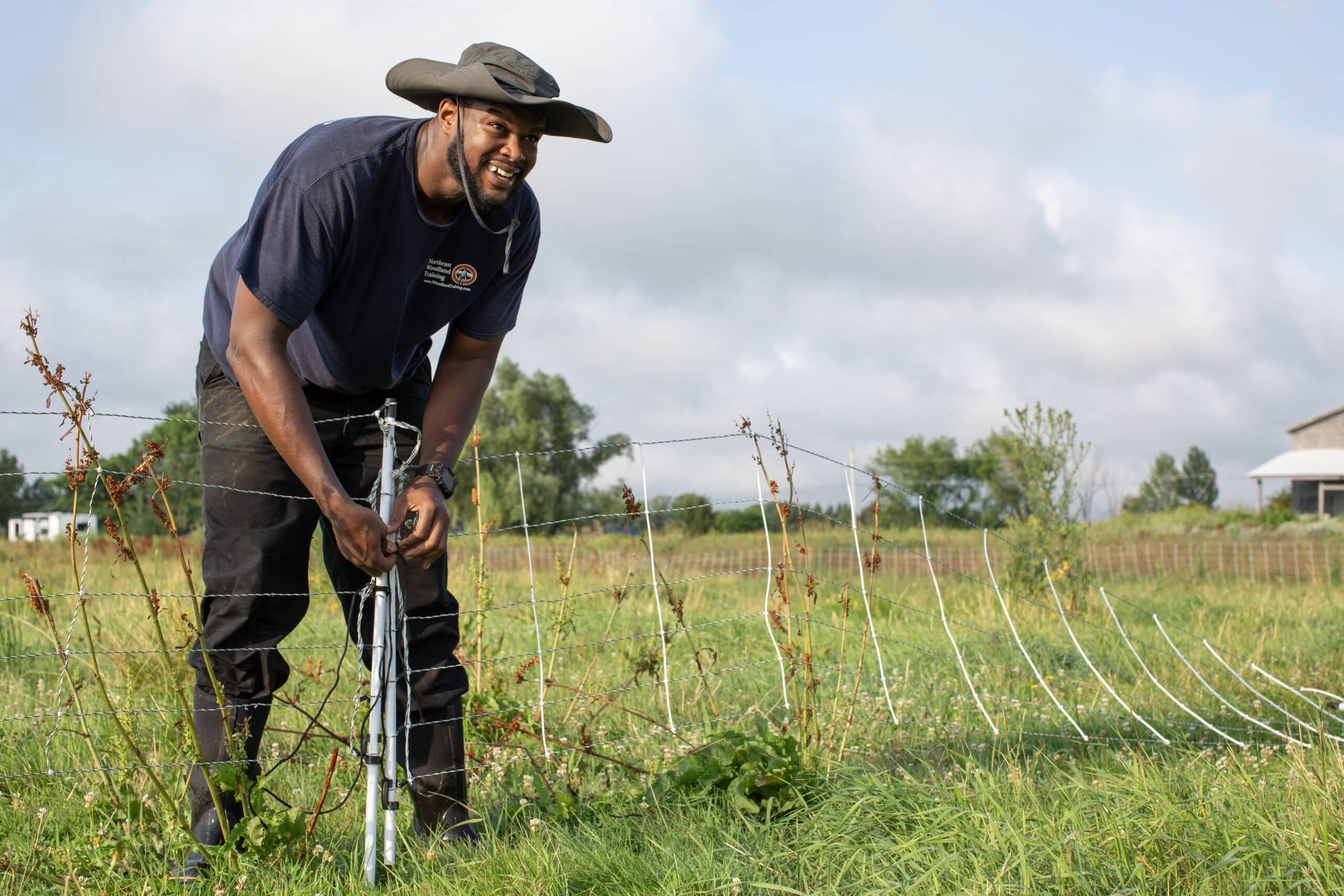 A man in a wide-brimmed hat and dark shirt works outdoors, repairing or setting up a wire fence in a grassy field under a partly cloudy sky. Shrubs and a house are visible in the background.