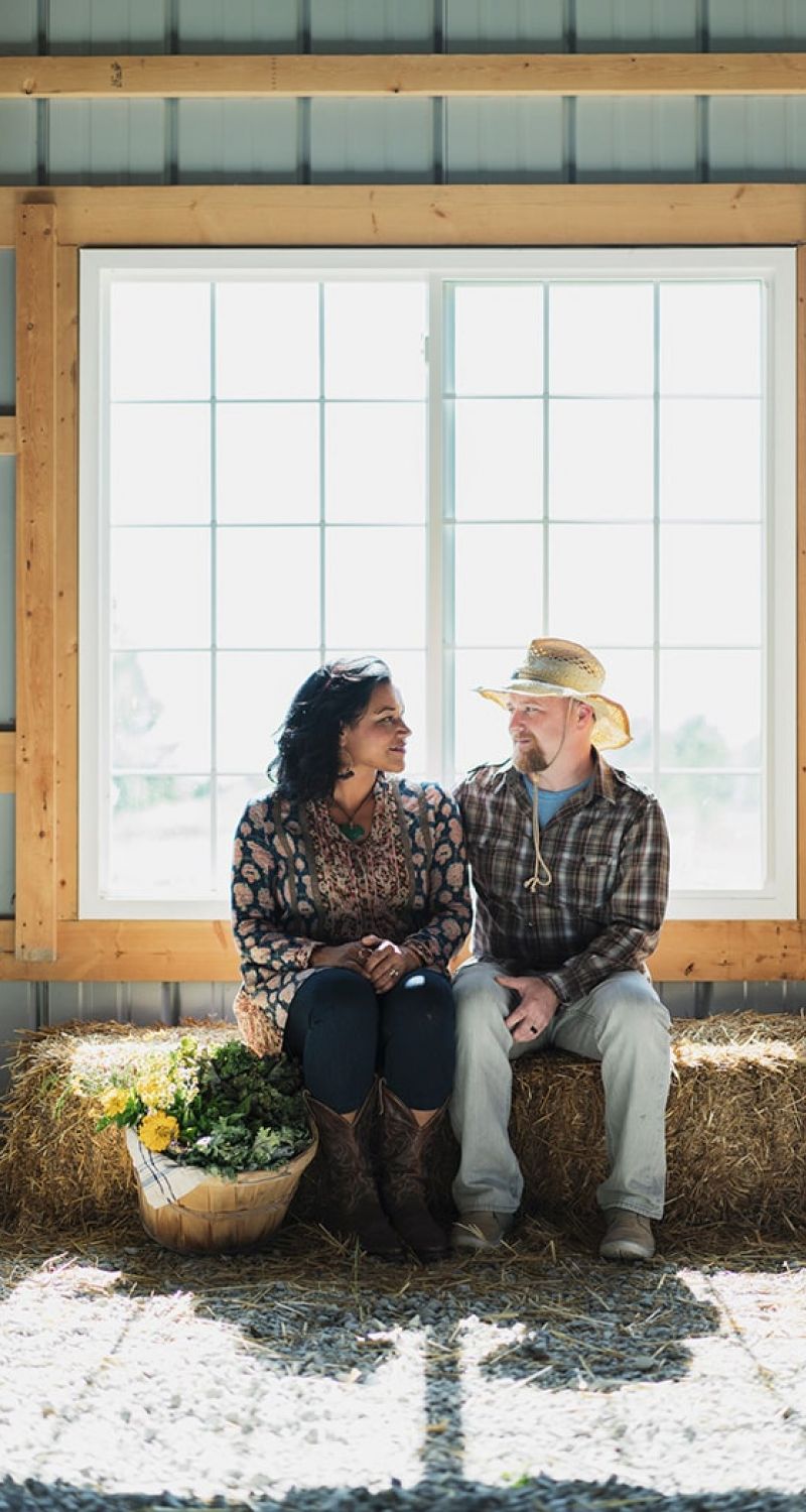 A woman and a man sit on a hay bale inside a barn, smiling at each other. The man wears a straw hat and plaid shirt; the woman wears patterned clothing. A basket of vegetables sits nearby, sunlight streaming through a large window behind them.