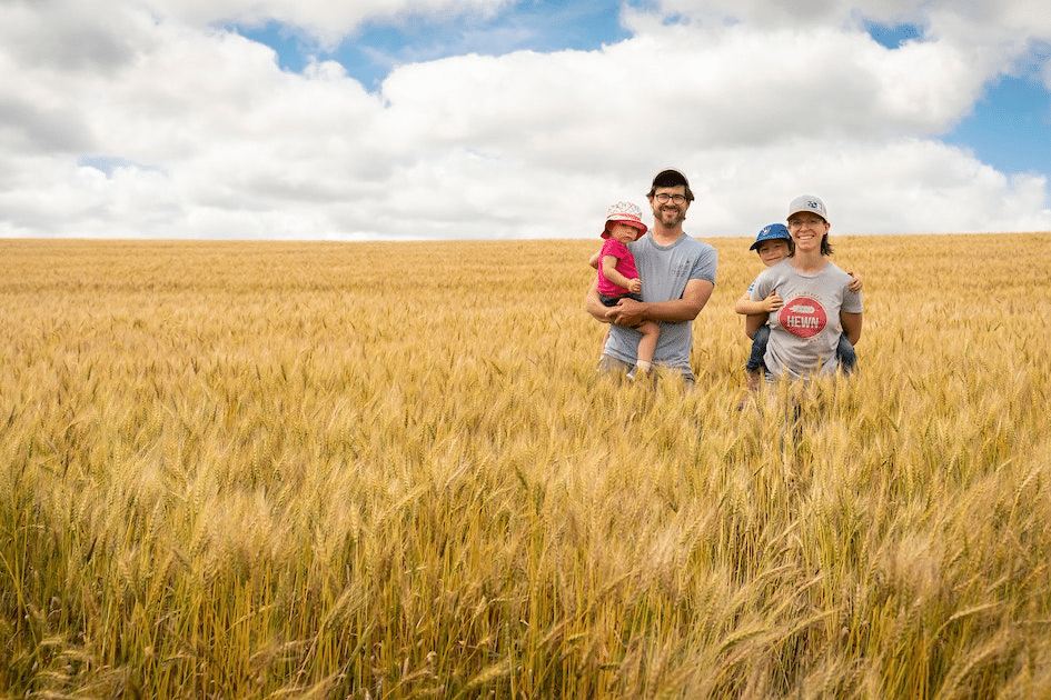 A family of four stands in a golden, organic wheat field under a cloudy sky. The father carries a small child in pink, while the mother holds a toddler in blue. Both parents wear casual clothes and baseball caps, smiling at the camera, embodying the joy of regenerative farming.