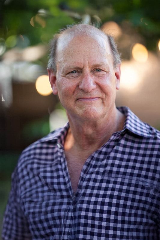 An older man with short, thinning hair smiles gently while wearing a checkered shirt. The background is softly blurred with green hues and round lights, suggesting an outdoor setting.