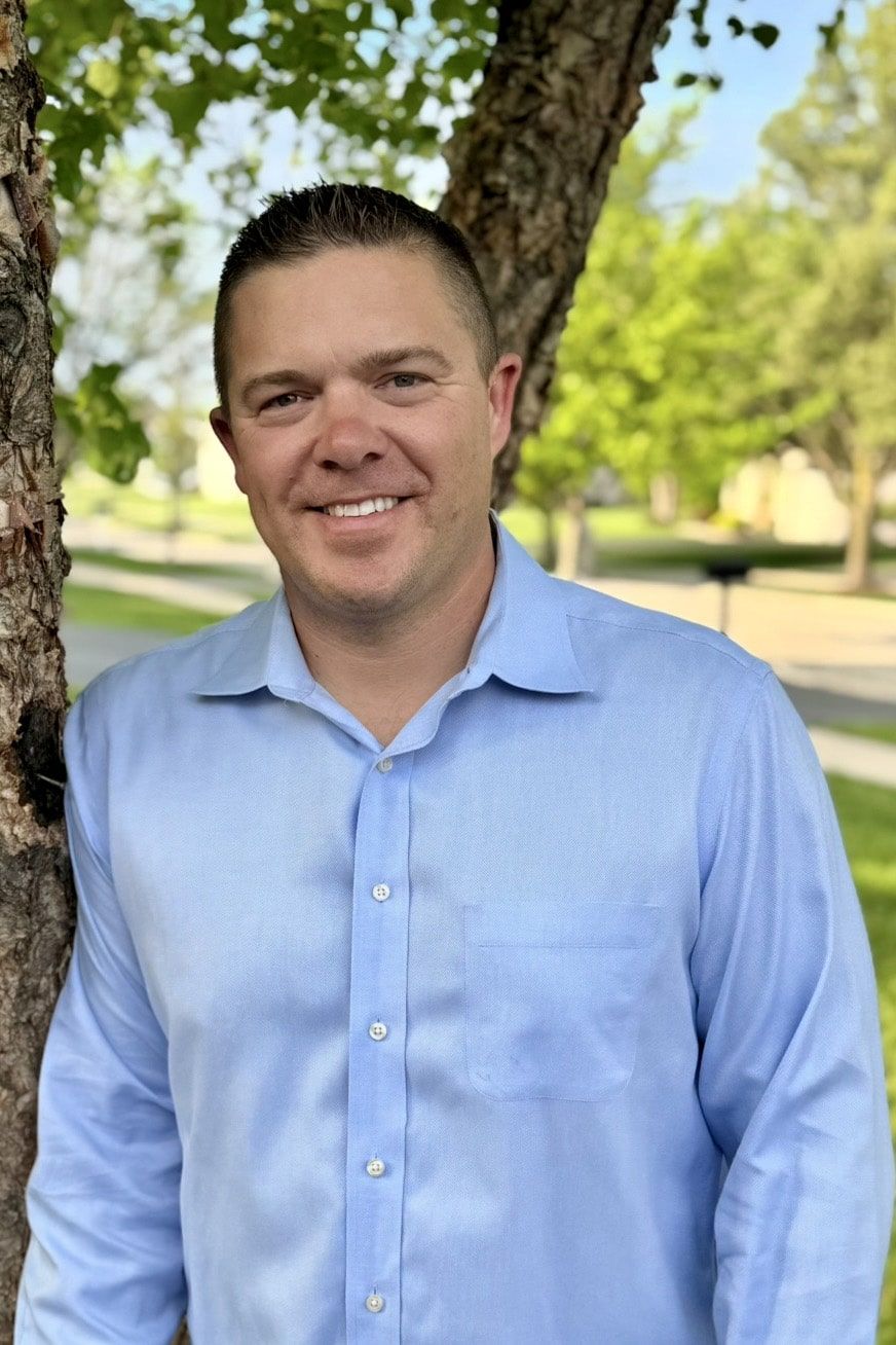 A man in a light blue button-up shirt smiles while standing outdoors, leaning against a tree with green leaves. The background features grass, trees, and a sunny street scene.