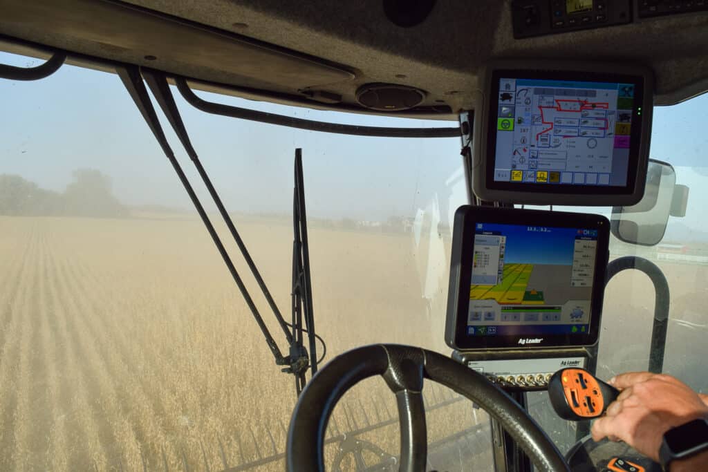 Inside the cabin of a combine harvester, the steering wheel, joystick, and digital screens display vital agricultural data. Through the window, a field of organic crops stretches out, highlighting ongoing regenerative harvesting activity.