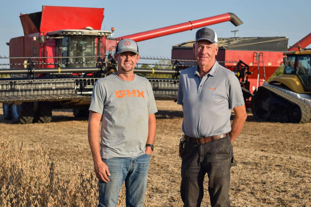 Two men in casual attire stand in a field, embodying the spirit of organic farming, in front of a red combine harvester and a tractor. The man on the left wears a grey shirt and cap, while the man on the right sports a polo shirt and cap. The field sets an undeniably regenerative tone.