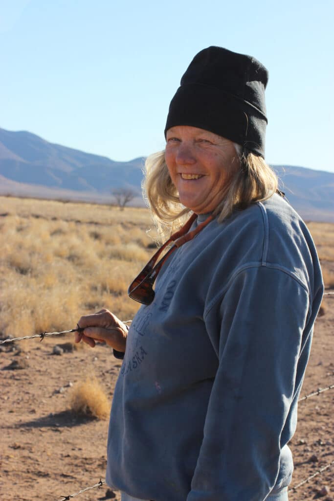 A woman wearing a black beanie and blue sweatshirt smiles while standing in a dry, open landscape with mountains in the background. She is holding a barbed wire fence.