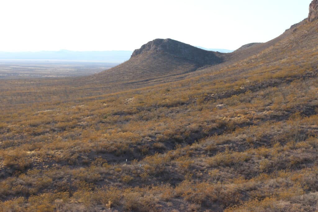 A vast arid landscape featuring rolling hills and sparse vegetation under clear skies. A prominent rocky hill rises in the middle distance, with mountains faintly visible on the horizon. The terrain is covered with dry shrubs and grasses.