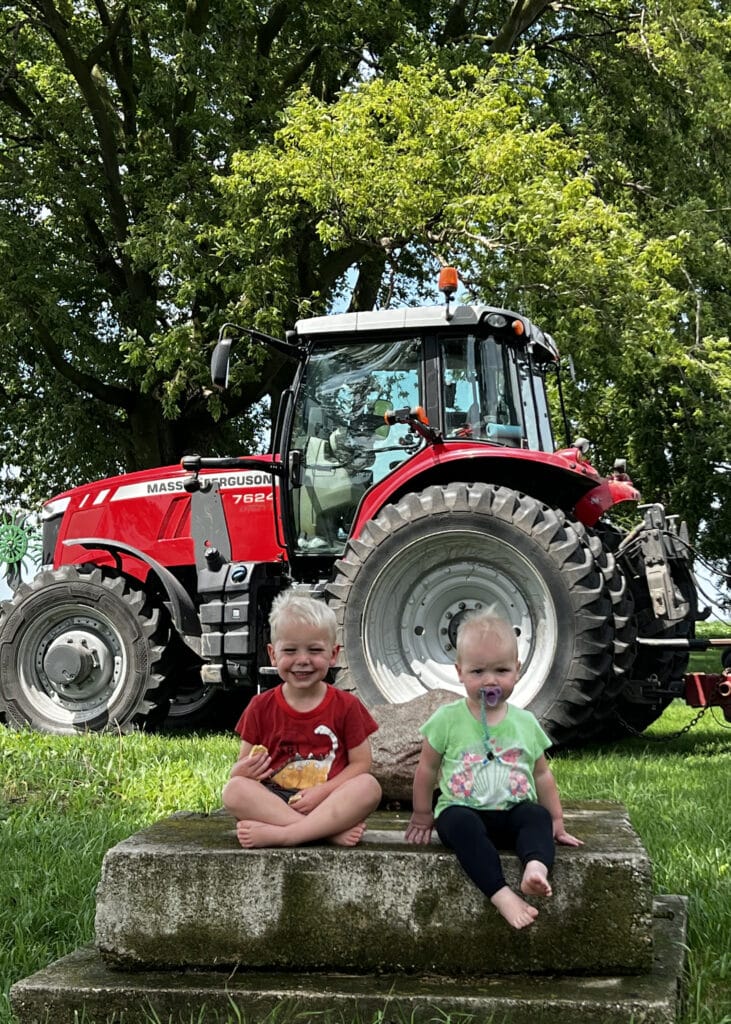 Two young children sit barefoot on a stone step in front of a large red tractor, with green grass and a leafy tree in the background. The children are smiling, and one has a pacifier.