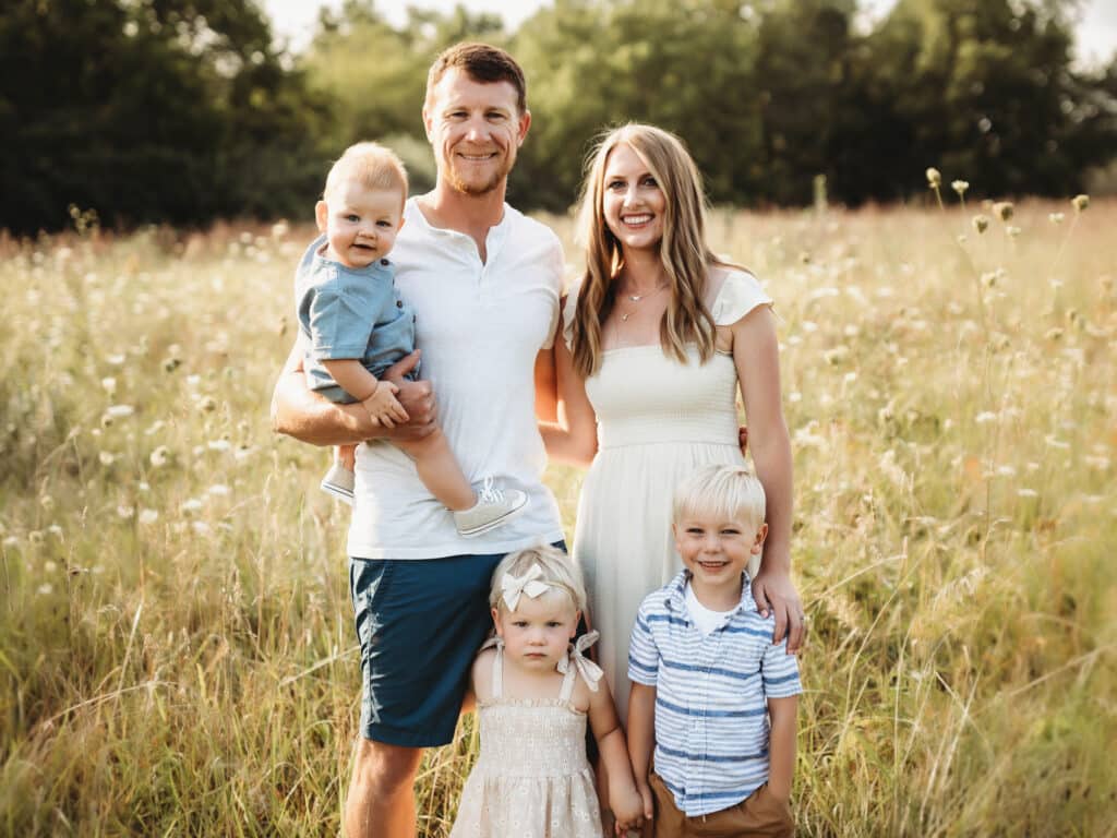 A smiling family of five poses outdoors in a sunlit field. Two adults stand behind their three young children, one of whom is held by the father. The grass is tall and green, with trees in the background.