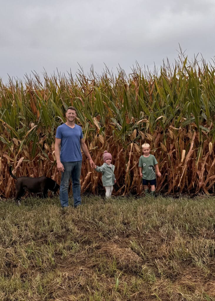 A man stands in front of a tall cornfield holding hands with a young child; another child stands nearby. A black dog sniffs the ground beside them. The sky is overcast and the grass is patchy.