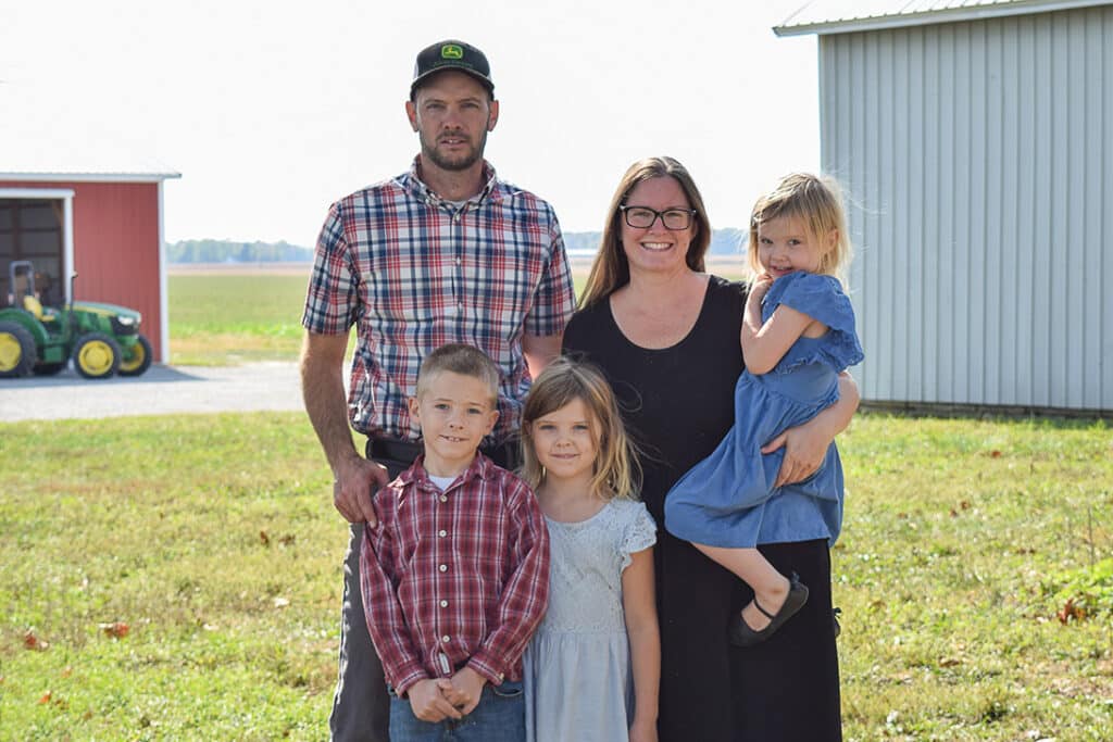 A family of five stands outside on grass near farm buildings and a green tractor. The man wears a plaid shirt and cap, the woman wears glasses and black dress, and three children—a boy and two girls—stand in front, one girl held by the woman.