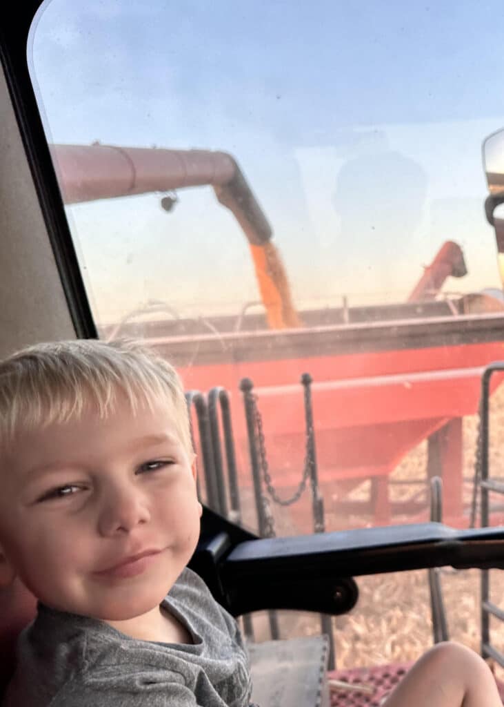 A young boy sits inside a vehicle, smiling at the camera. Outside the window, corn is being loaded from a combine into a red grain cart in a field during harvest.