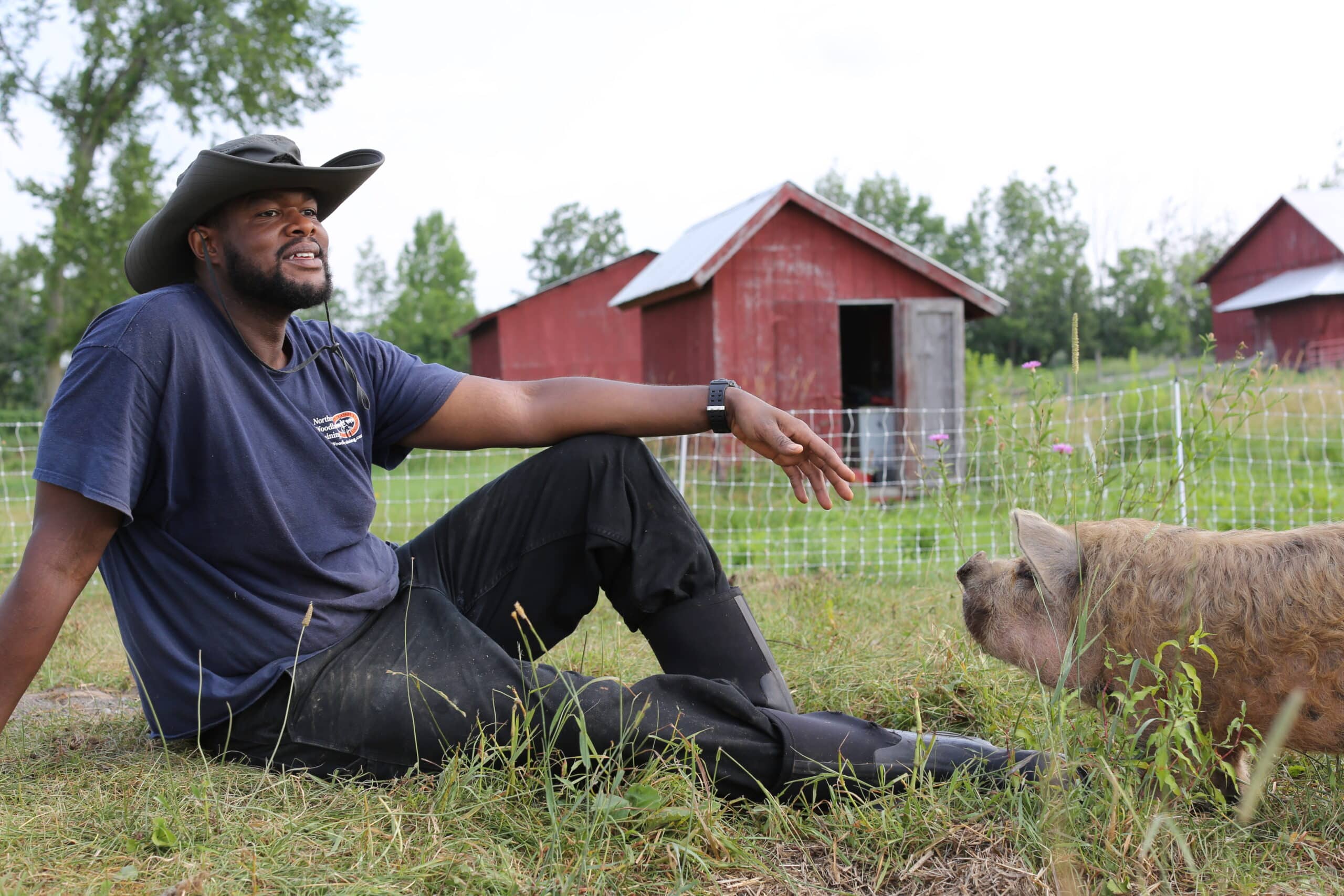 A man wearing a wide-brimmed hat and boots sits on the grass, smiling at a pig nearby. Red barns and a wire fence are visible in the background on a sunny day.