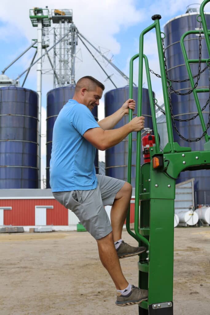 A man in a light blue shirt and gray shorts climbs green metal equipment on a farm, with large blue silos and a red barn in the background under a partly cloudy sky.