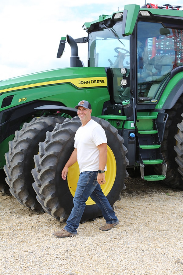A man in a white t-shirt, jeans, and a cap walks in front of a large green John Deere tractor with dual rear wheels on a gravel surface.