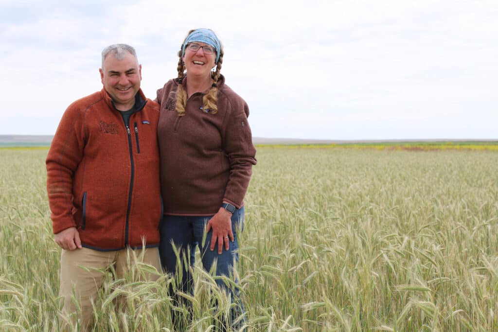 Two people stand smiling in a field of tall green grass. Both wear brown jackets; one person has braided hair and a blue headband. The sky is overcast, and the landscape is open and rural.