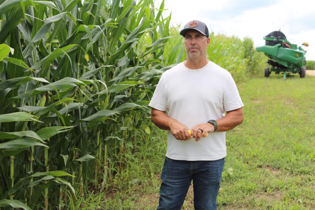 A man in a white shirt and jeans stands by tall corn plants in a field, holding corn. A green combine harvester is visible in the background on a grassy path.