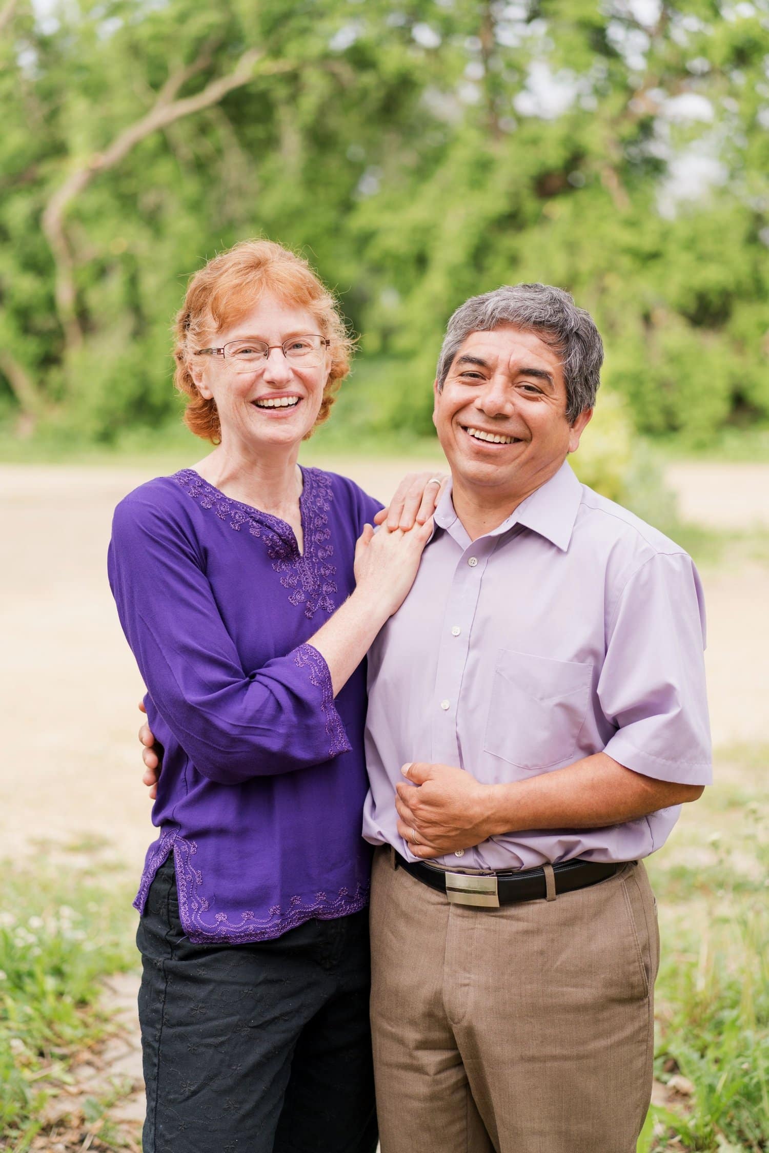 A smiling couple stands outdoors, both wearing purple tops. The woman has red hair and glasses, and her hand rests on the man's shoulder. The man has short gray hair. Green trees and a bright background are visible behind them.