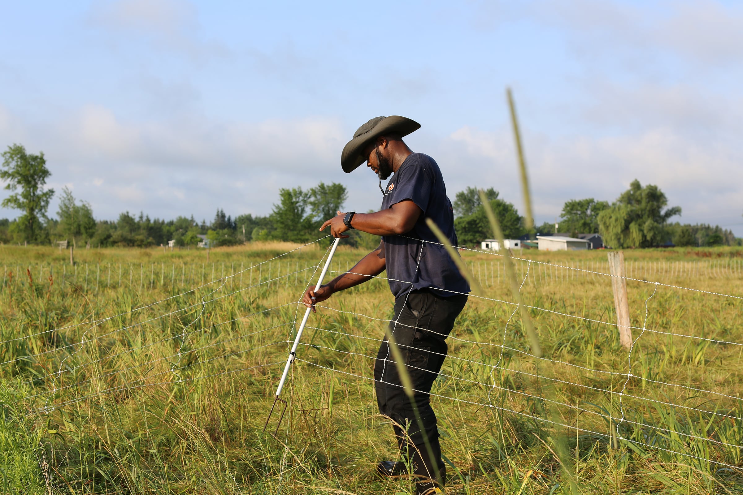 A person wearing a wide-brimmed hat and dark clothing installs or repairs a wire fence in a grassy field under a partly cloudy sky, with trees and buildings visible in the background.