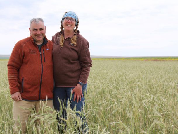 Two people stand smiling in a field of tall green grass. Both wear brown jackets; one person has braided hair and a blue headband. The sky is overcast, and the landscape is open and rural.