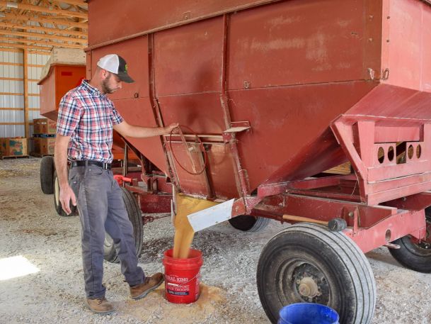 A man in a plaid shirt and cap stands next to a large red grain cart, filling a red bucket with grain inside a barn. The grain is pouring from a chute at the bottom of the cart.