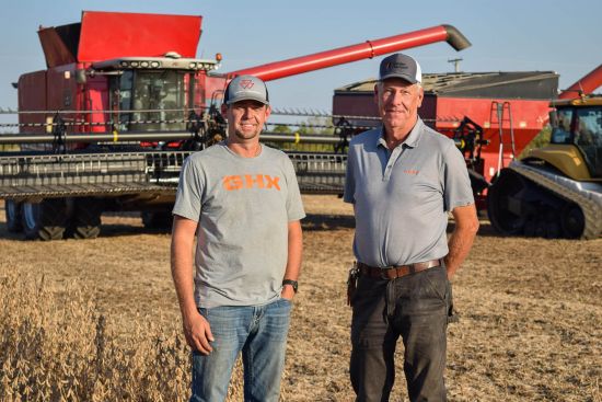 Two men stand in a harvested field with a red combine harvester and other farming equipment in the background. Both wear caps and gray shirts, and they are smiling at the camera on a sunny day.